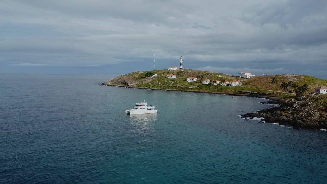 Catamarano di supporto per immersioni nel Parco Nazionale Marino di Abrolhos con il Farol de Abrolhos sull'isola di Santa Bárbara, Caravelas Bahia Brasile, ripresa aerea con drone in allontanamento