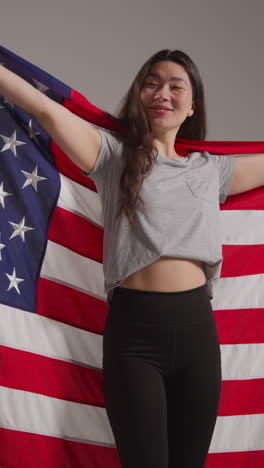 Toma-De-Estudio-De-Video-Vertical-De-Una-Mujer-Sosteniendo-Una-Bandera-Estadounidense-Detrás-De-Ella-Celebrando-El-4-De-Julio,-Día-De-La-Independencia.