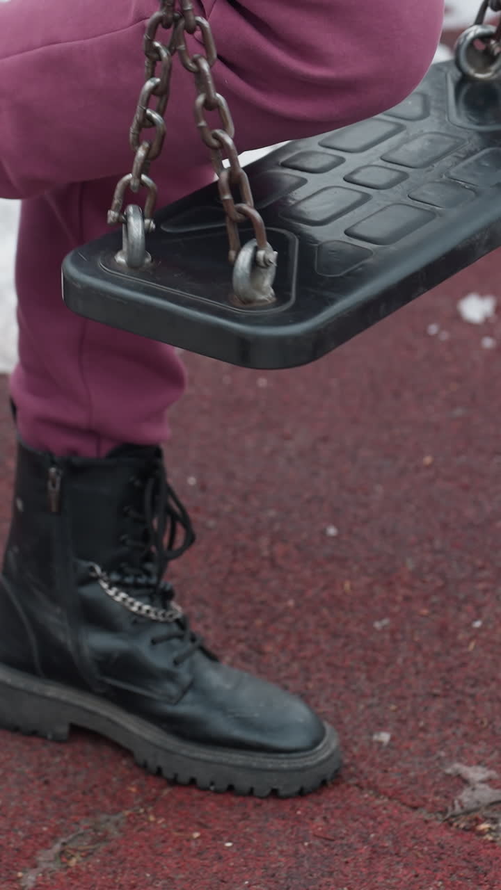 vista de la pierna de una persona con botas negras arrodillada en un columpio de hierro sobre un suelo de goma roja con manchas de nieve, entorno de parque de invierno al aire libre con atmósfera de clima frío y una mezcla de texturas