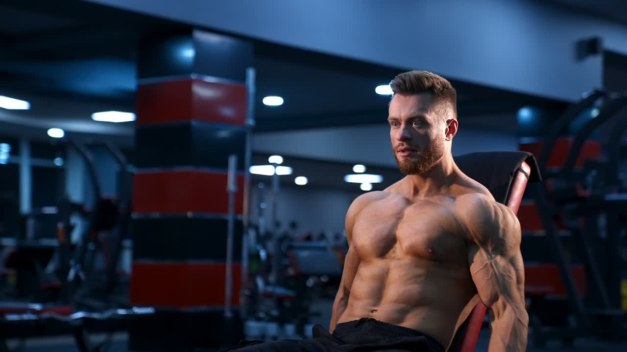 Athletic handsome man trains with dumbbells on fitness chair. Modern gym background. Slow motion.
