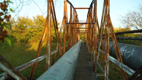 Durchgang-Durch-Eine-Alte-Industrielle-Rostige-Eisenbrücke-Mit-Vielen-Rohren,-Die-Dampf-Und-Heißes-Wasser-über-Einen-Schmalen-Fluss-An-Einem-Sonnigen-Tag-Leiten,-Aufgenommen-In-Zeitlupe-Mit-120-Fps