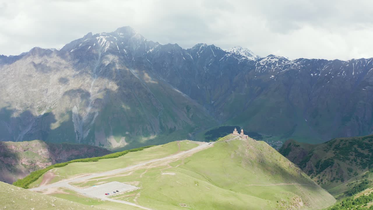 Ripresa aerea di presentazione con drone della cattedrale della Santissima Trinità di Gergeti su una collina, con un'imponente catena montuosa sullo sfondo.