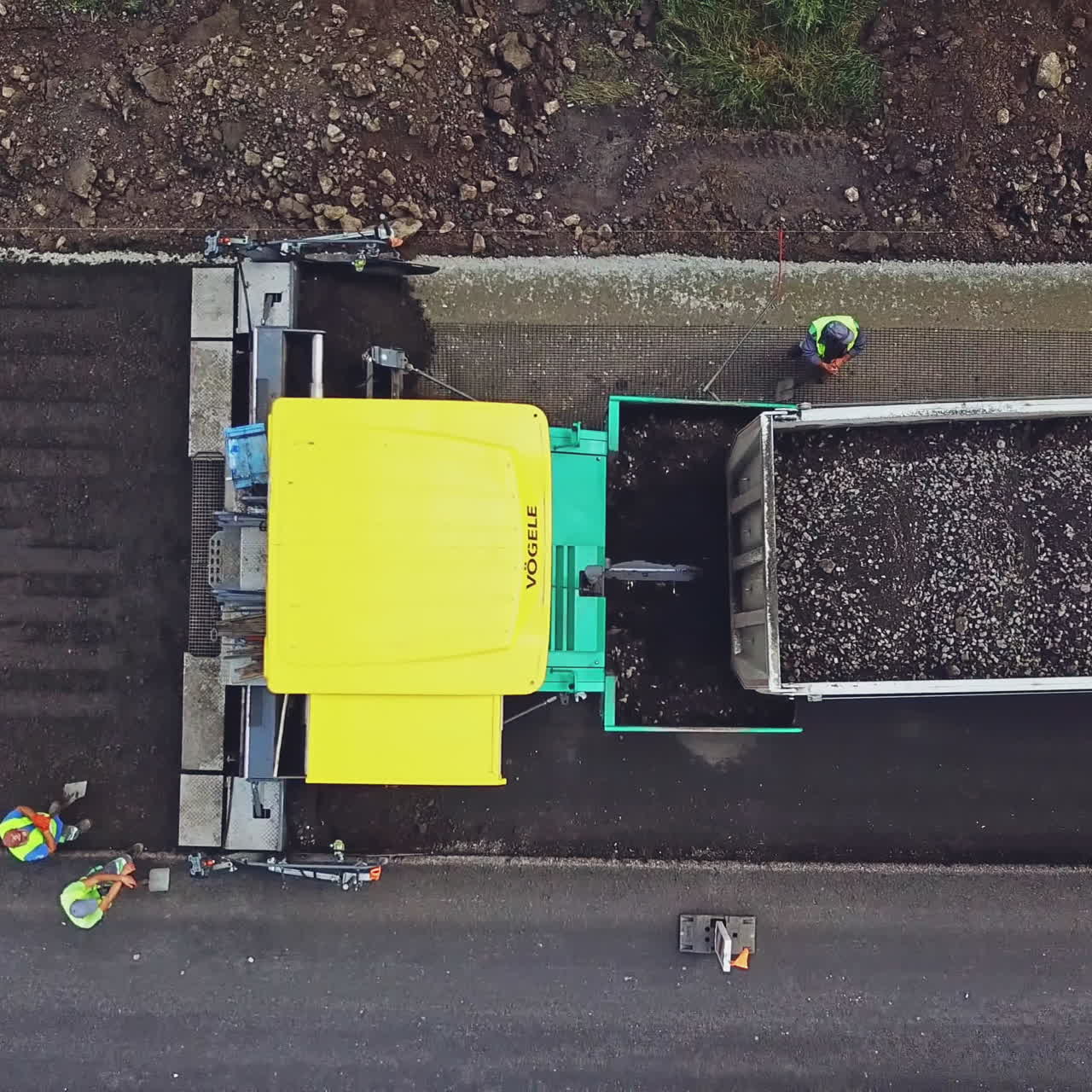 A big truck with bitumen and rubble pulls up to the paver on the background of a fresh asphalt strip and a few people around. Road repair. Aerial view.