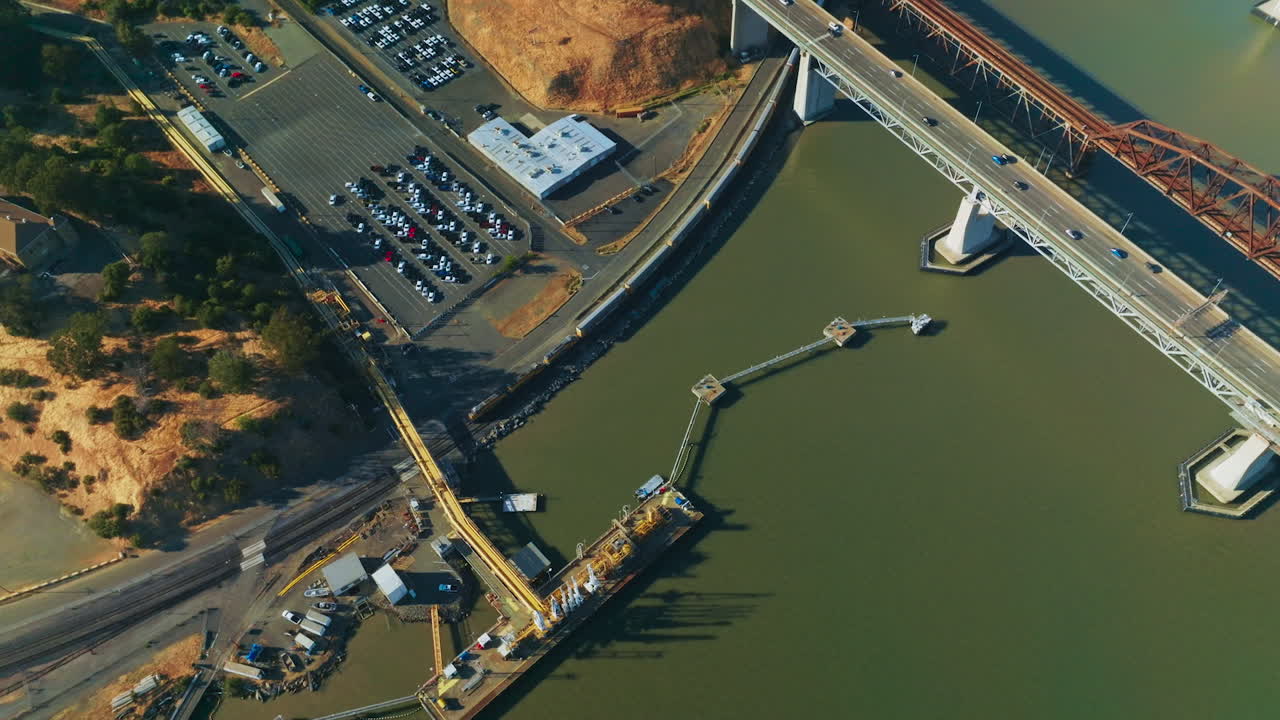 Train moving at the waterfront of California bay under Martinez bridge. Industrial area with docks and big parking lots. Top view.
