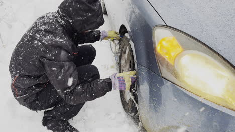 Man-installing-snow-chains-on-the-wheels-of-car,-winter-landscape-with-falling-snow,-on-road-full-of-snow,-side-view