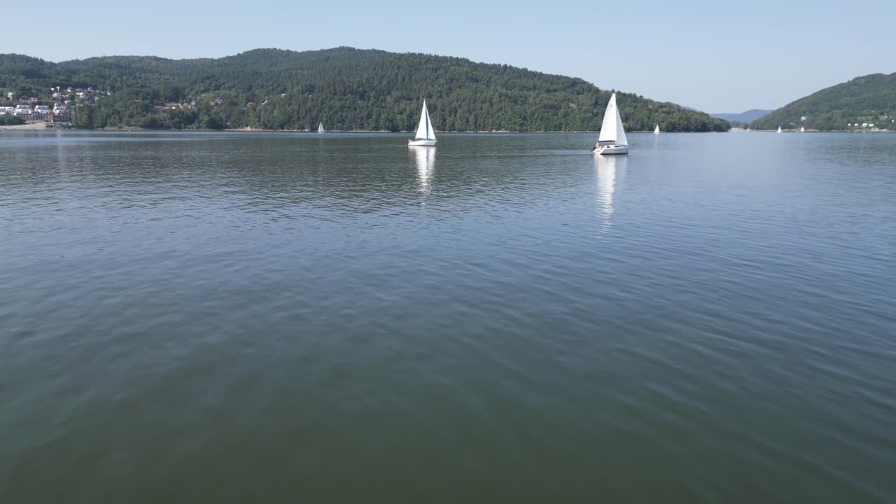 barcos de vela blancos en el lago pacífico en las montañas beskid, rodeados de vegetación durante un día de verano - vista aérea 4k