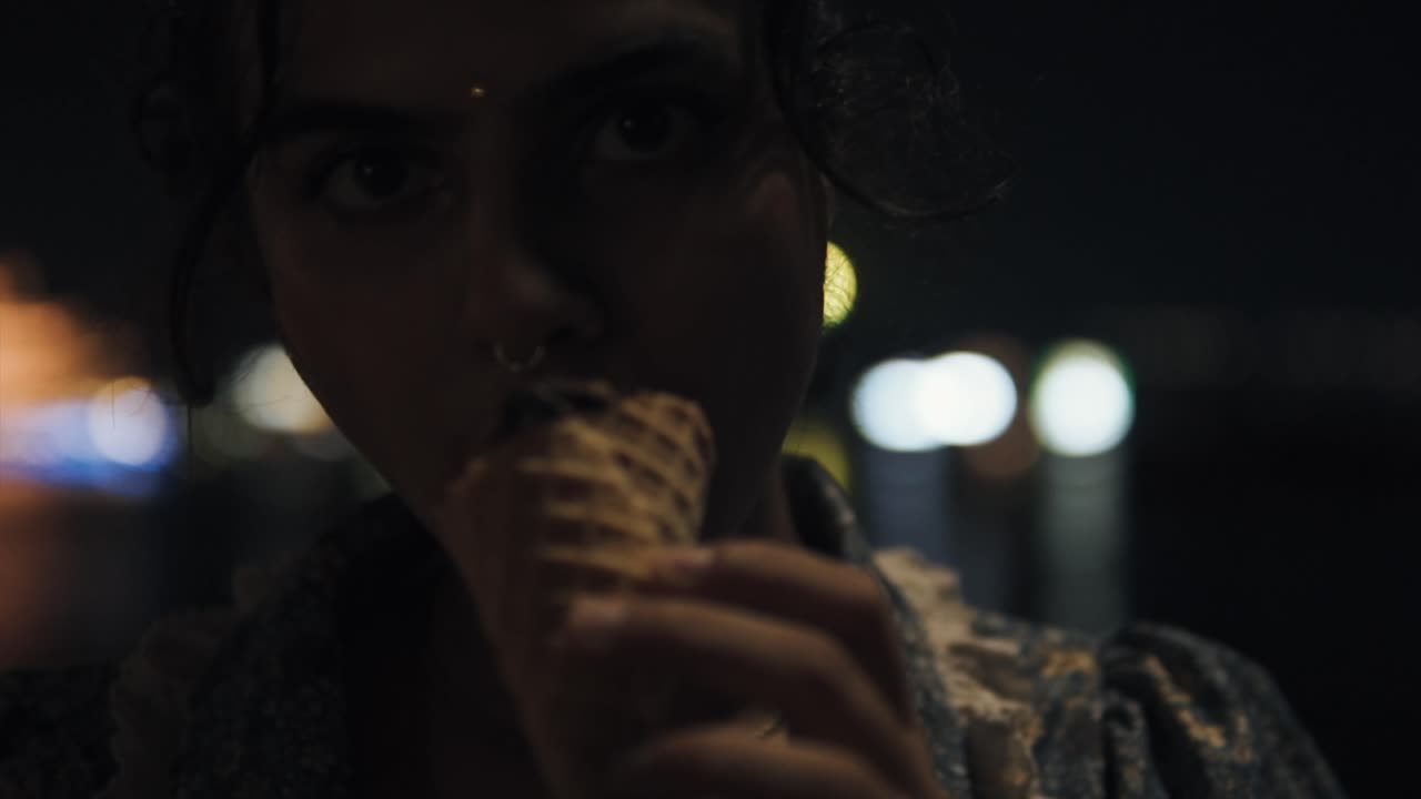 mirando fijamente a la cámara de una chica india lamiendo un cono de helado por la noche en la ciudad