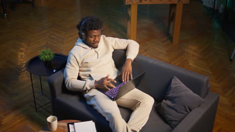 african american self employed man browses the web and listens to music, having fun with songs on headset at his modern home. young adult laying on couch and relaxing in the living room. camera b.