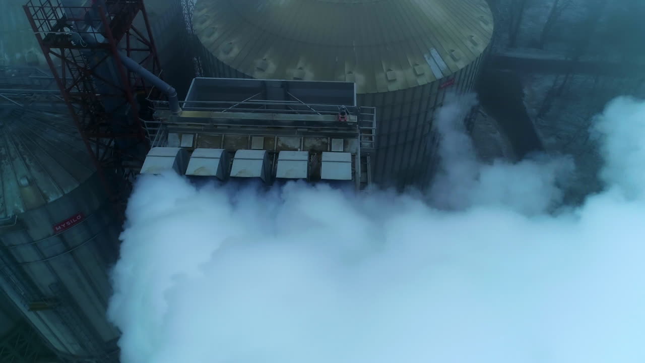 Five connected pipes producing heavy thick white smoke. Granary complex premises and tanks from aerial perspective on grey winter day.