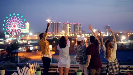 happy friends are burning bengal lights and dancing standing on rooftop celebrating holiday together. entertainment, friendship, big city and joy concept.