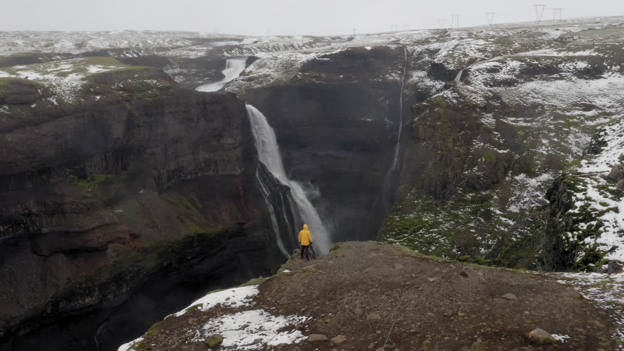 antena: toma panorámica lenta de un hombre parado cerca del borde de un acantilado, entre las cascadas haifoss y granni en islandia