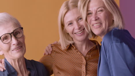 two blonde mature women and blonde senior woman smiling and hugging posing on orange and purple background