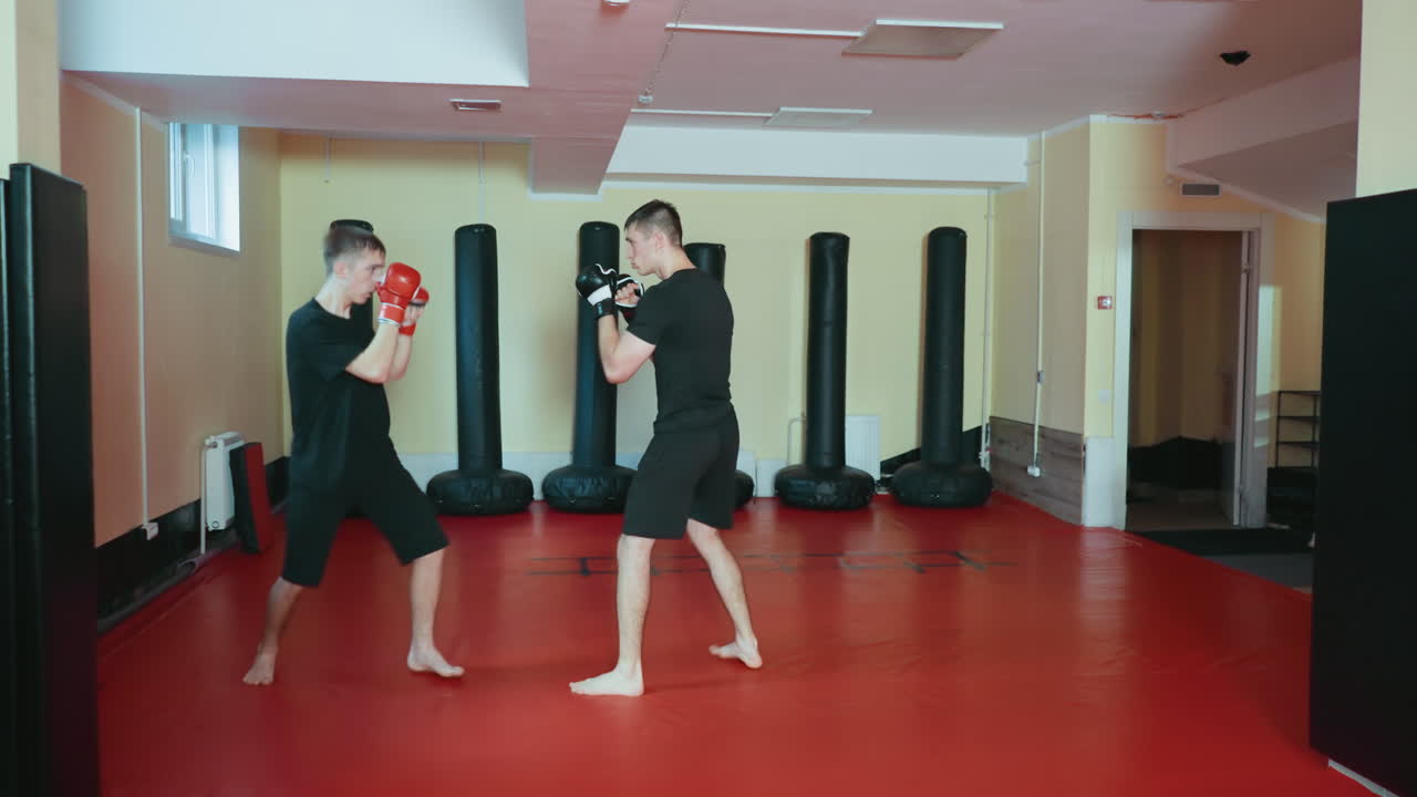 격투기 체육관 안에서 마주 보고 스파링을 준비하는 운동선수들의 모습이에요. 한 명은 빨간색 글러브를 들고 있고, 다른 한 명은 검은색 글러브를 끼고 방어 자세를 취하고 있어요. 두 선수 모두 맨발로 빨간색 훈련 매트 위에 서 있고, 근처에는 샌드백이 있어요.