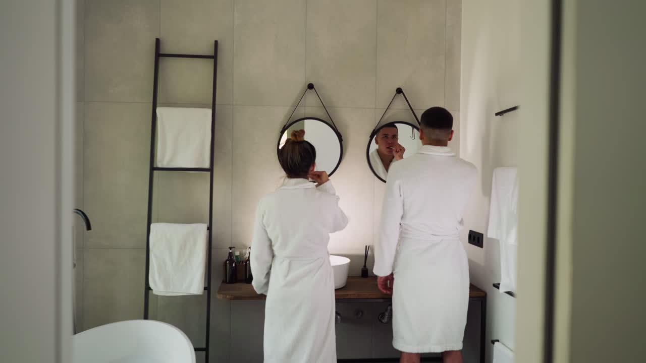 Lovers brush teeth in front of mirrors in bathroom. Couple in soft terry bathrobes performs everyday care procedures for dental health together