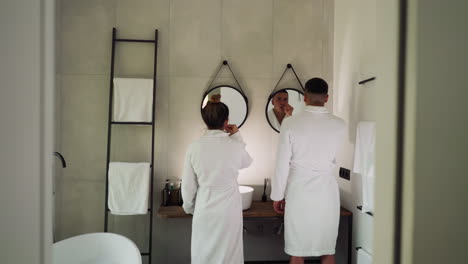 lovers brush teeth in front of mirrors in bathroom. couple in soft terry bathrobes performs everyday care procedures for dental health together