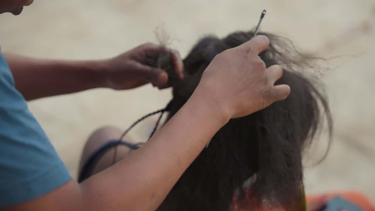 Mani che districano capelli naturali spessi. Un'intrecciatrice mobile scioglie nodi stretti con la punta delle dita, tirando con attenzione per prevenire la rottura. Terreno sabbioso, cliente seduta, ritmo del trattamento, texture resistente.