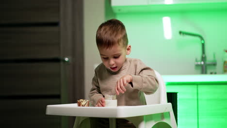 beautiful toddler boy sits at high chair in the kitchen. kid has a smack with yogurt and bun.
