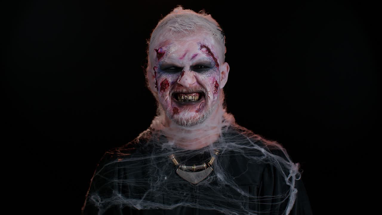 Zombie man with wounds scars and contact lenses looking at camera clicks his teeth trying to scare
