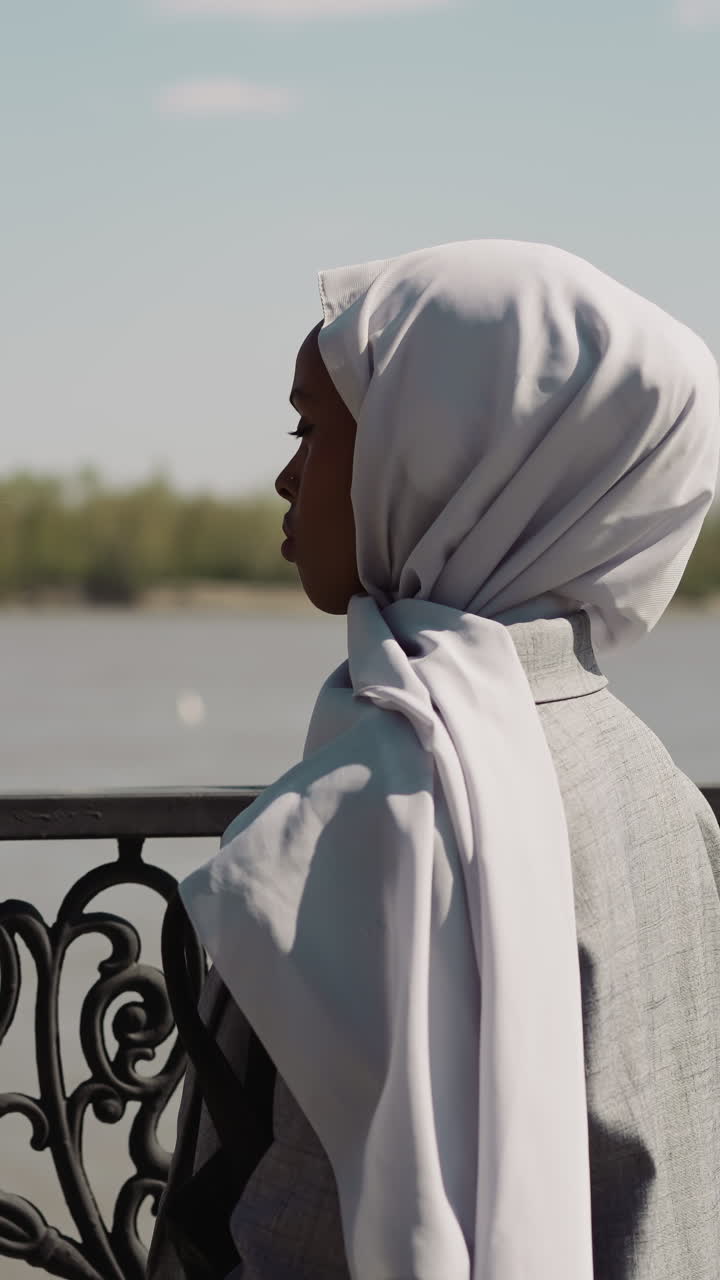 Thoughtful Muslim lady with chiffon hijab looks at distance near fence on embankment. African-American lady tourist in city. Travelling and vacation