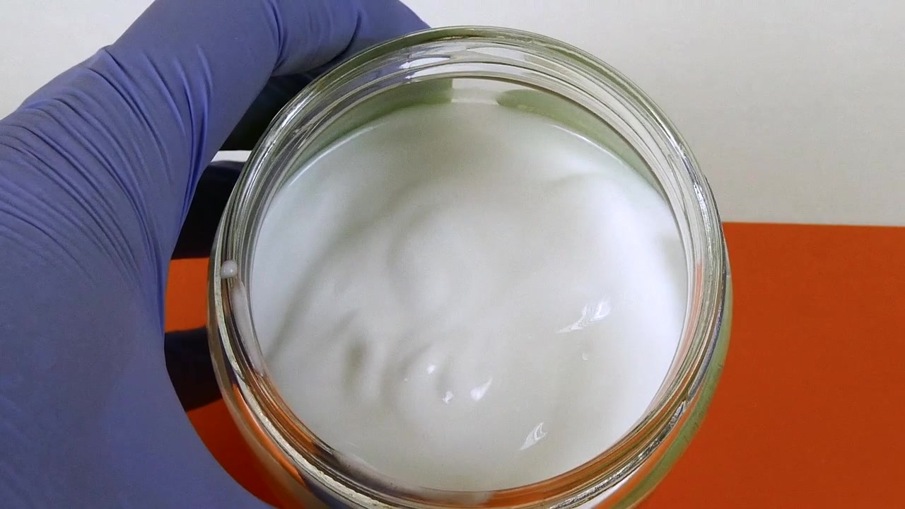 Mano professionale con un guanto blu che tiene un barattolo di vetro con crema bianca per test dermatologici.