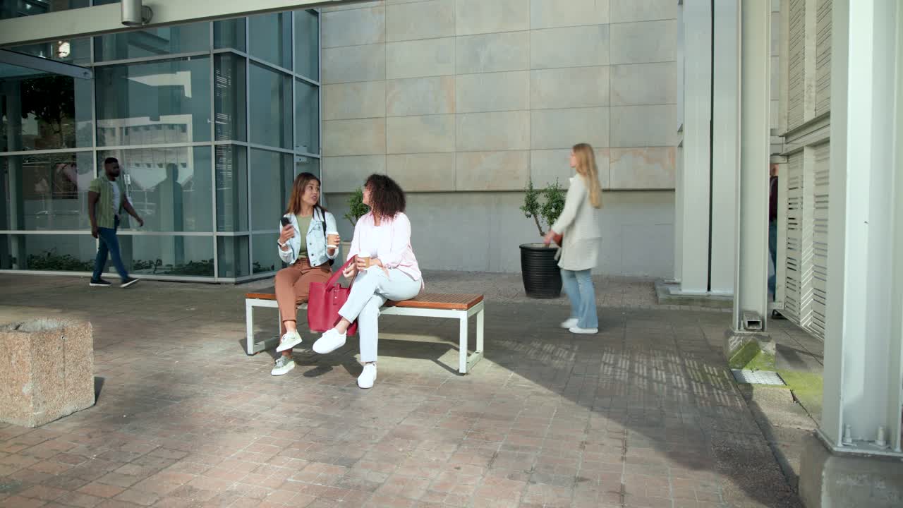 Due donne afroamericane che entrano da destra, si incontrano su una panchina, si siedono, chiacchierano, tenendo il caffè.