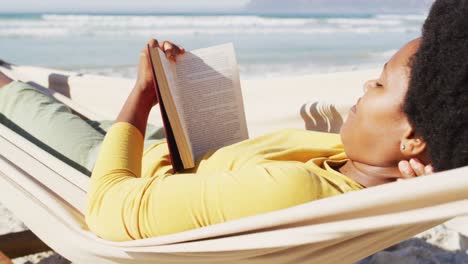 feliz mujer afroamericana leyendo y acostada en una hamaca en una playa soleada