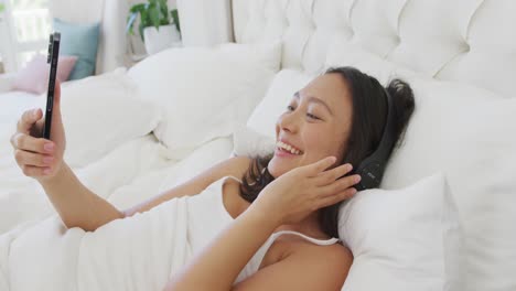 Glückliche-Asiatische-Frau,-Die-Im-Bett-Liegt,-Kopfhörer-Trägt-Und-Ihr-Smartphone-Benutzt,-In-Zeitlupe