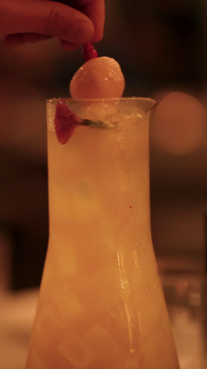 Una mano posiziona una guarnizione di litchi su un cocktail in un'illuminazione fioca e calda, creando un'atmosfera sofisticata
