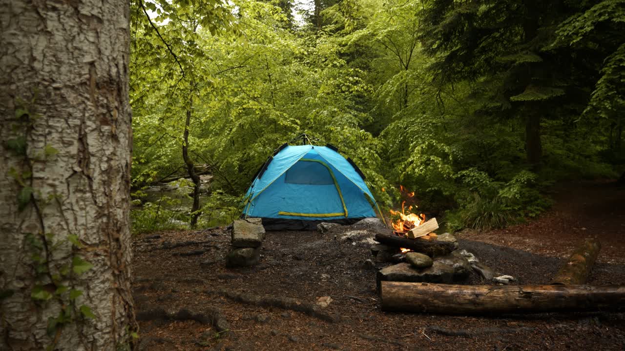 A fire burns at a campfire in the forest. A tent stands next to the burning campfire. It's raining, and the atmosphere is mystical. Shot in slow motion. A calming atmosphere without people.