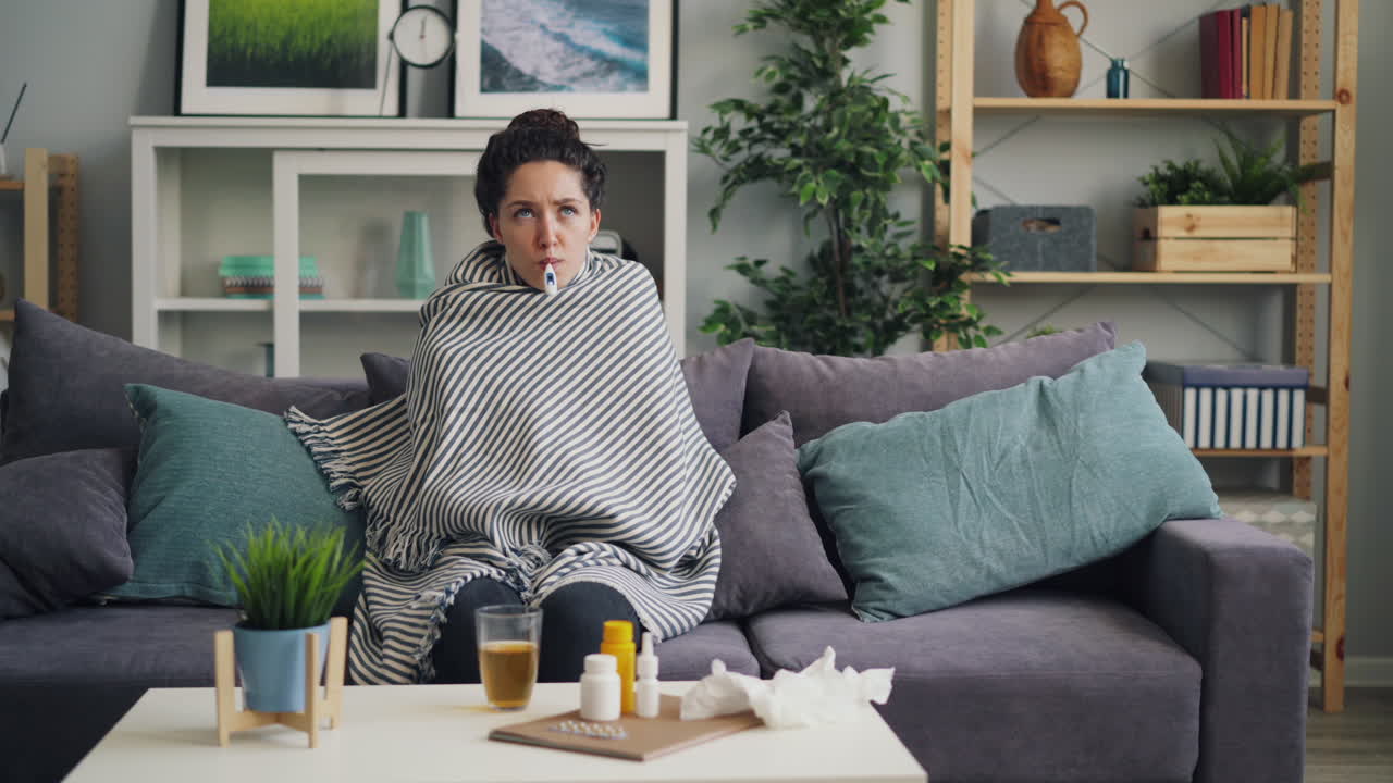 Woman resting on sofa with blanket, taking temperature