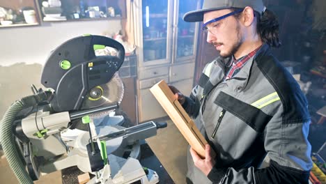 concentrated young bearded joiner in goggles and a cap checks the work piece