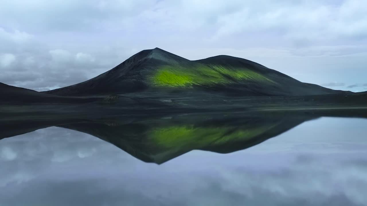 Aerial drone gliding over and up over a large reflective lake or water source with a large green moss and plants covered mountain in the background at Iceland or Greenland during a cloudy day.