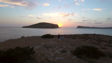 Toma-Aérea-De-Una-Pareja-En-Los-Acantilados-Viendo-La-Puesta-De-Sol-Sobre-El-Mar.