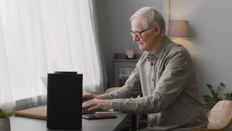 Glücklicher-älterer-Mann,-Der-Am-Laptop-Schreibt,-Während-Er-Am-Schreibtisch-Sitzt