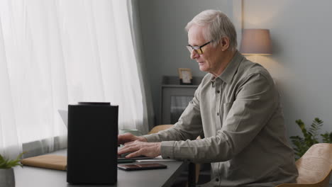 feliz anciano escribiendo en la computadora portátil mientras está sentado en el escritorio