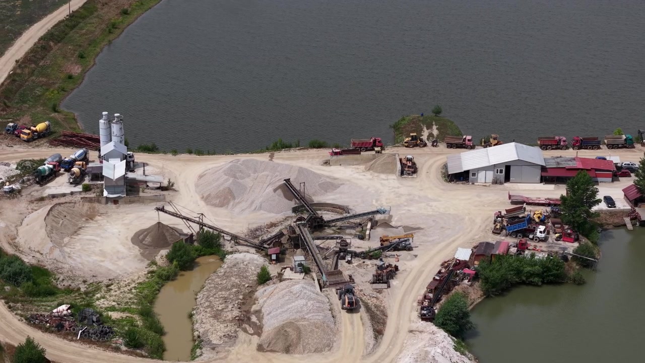 Aerial view shows an active industrial sand and gravel quarry, with heavy machinery processing aggregates next to a lake, crucial for construction.