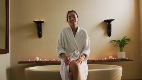 happy biracial woman sitting in robe on bathtube