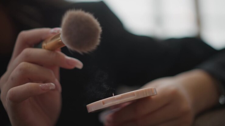 Tæt nærbillede fra siden af hænder, der holder en blød makeupbørste over en lyserød pudderdåse med synlig fejende bevægelse og pudder, der klæber til børstehårene, hvilket skaber en blød opfriskning med roligt indendørs lys