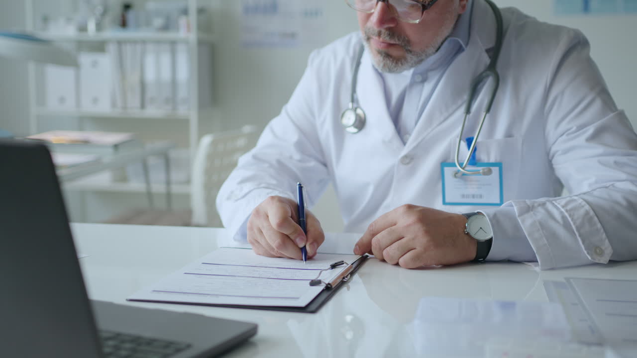 Doctor Reading Information on Laptop and Writing Medical Records at Desk