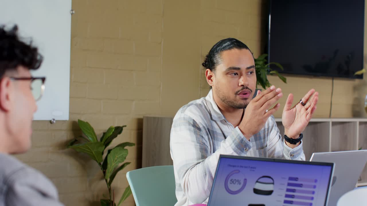 Mid adult man with bun leaning ahead gesturing spacing and explaining laptop charts in meeting room