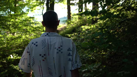 Young-man-walks-barefoot-in-slow-motion-out-of-a-dark-forrest-towards-a-bright-green-meadow-in-with-sunshine