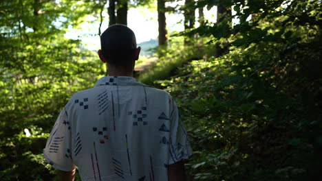 young man walks barefoot in slow motion out of a dark forrest towards a bright green meadow in with sunshine