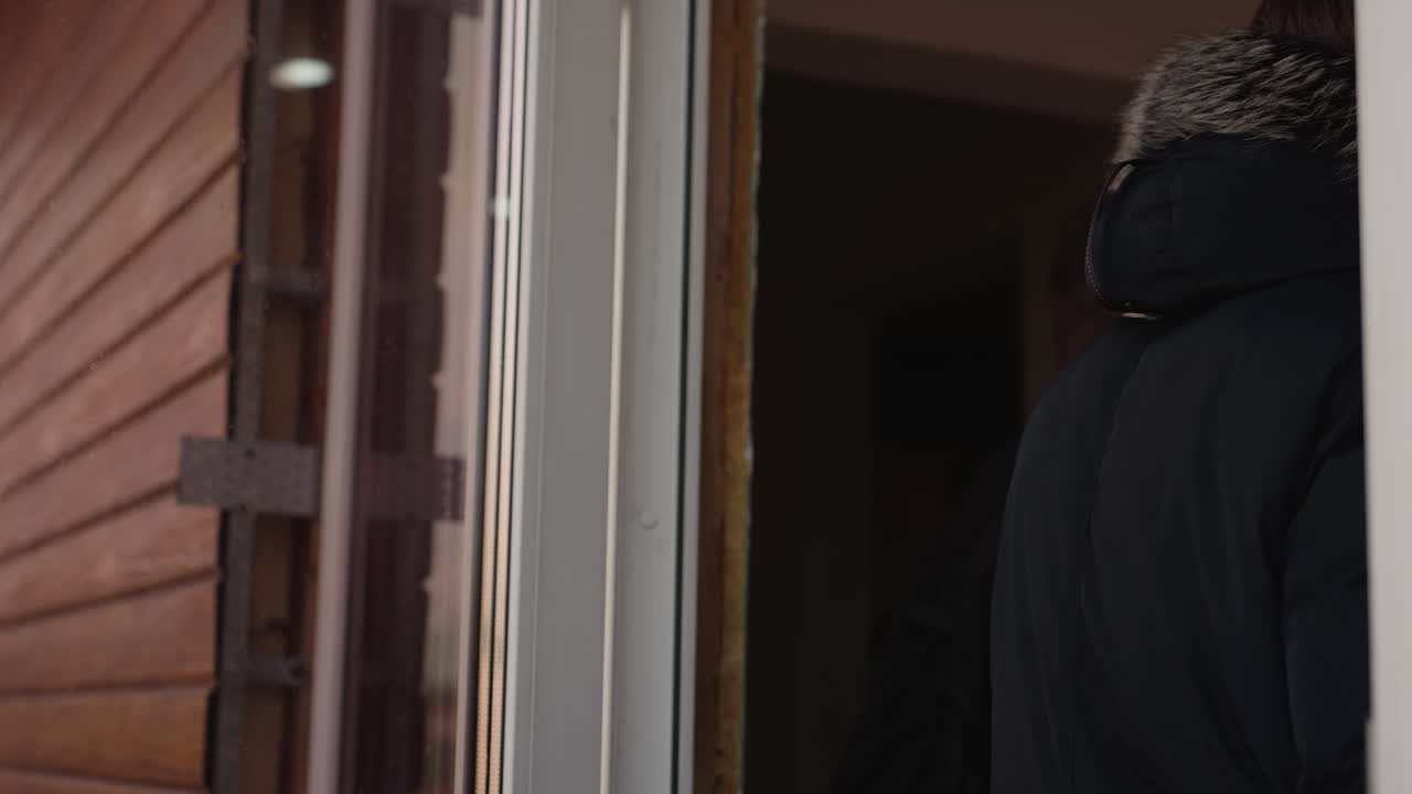 Young man with long hair wearing winter jacket with fur hood walks into a wooden building, side profile view, looking into distance, cold weather atmosphere, city street background, casual lifestyle