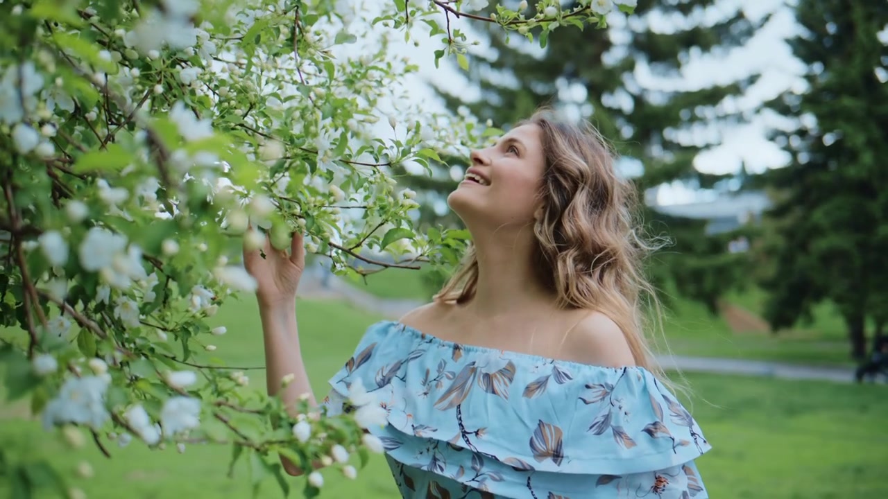 Giovane donna sorride dolcemente mentre tocca i rami fioriti di un albero. Il vento le solleva i capelli mentre si china verso i fiori primaverili. La scena cattura un momento di pace in un ambiente di parco verde.