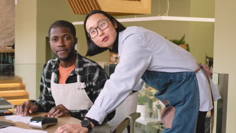 two multiethnic coffee shop owners smiling at camera while calculating finance bills on laptop computer