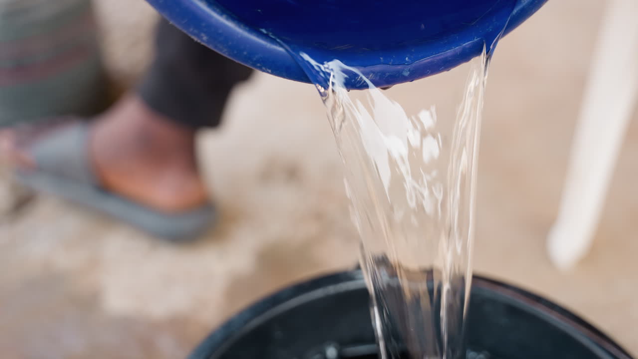 Operaio che versa acqua da un secchio blu, piedi con infradito visibili, flusso costante in un contenitore nero, acqua del cantiere per miscelazione di falegnameria e risciacquo attrezzi, pavimento in cemento esterno, primo piano di flusso e schizzi.