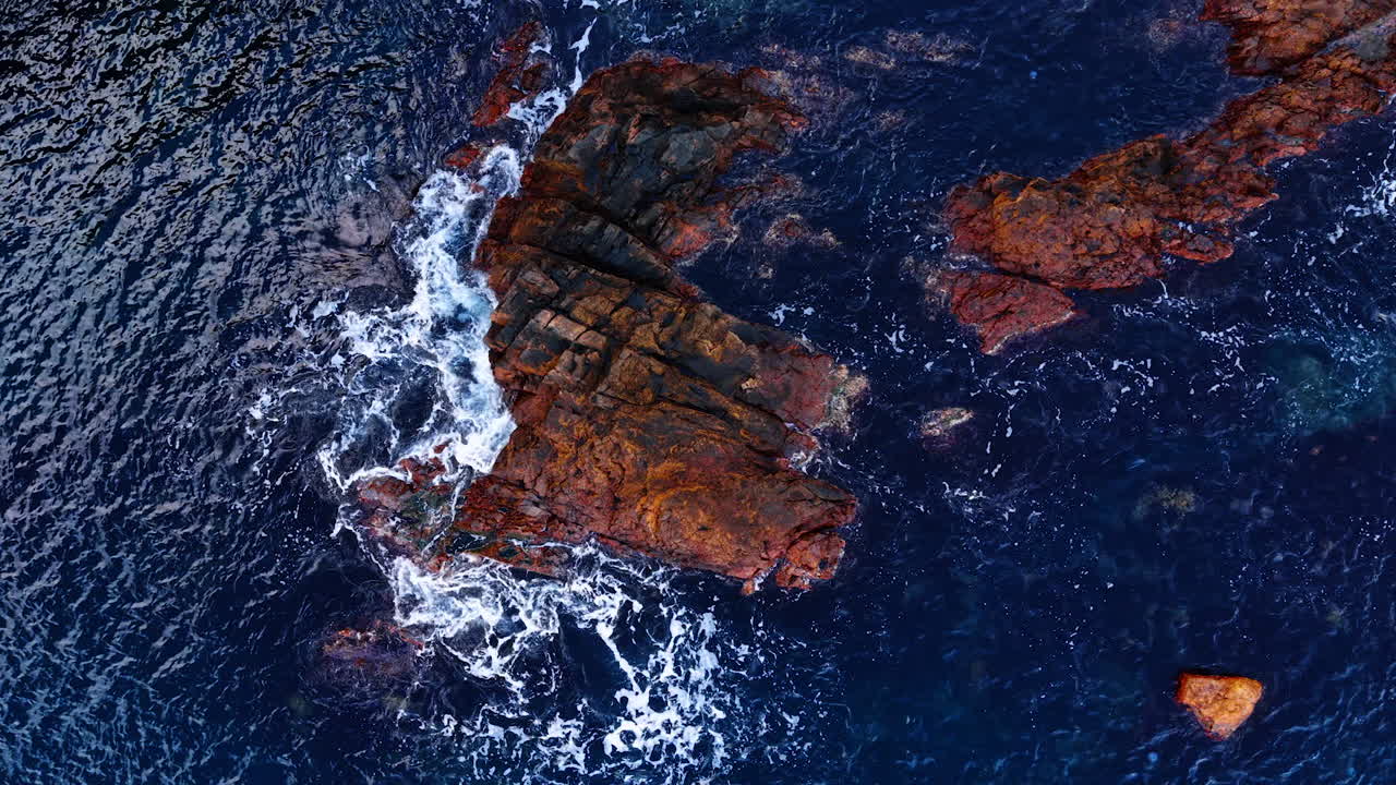 Aerial View of Rocky Coastline with Ocean Waves Splashing Against Rocks. Aerial shot captures dramatic ocean waves crashing against rugged, reddish-brown rocky formations in deep blue sea