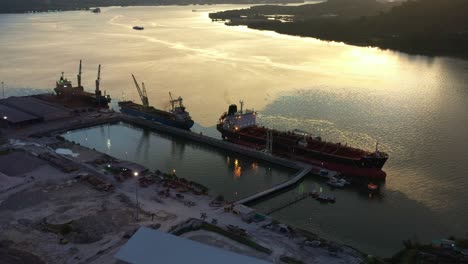 birds eye view fly around lumut port industrial park along sungai manjung river with commercial bulk carrier ships docked at the port, tilt up reveals mountain landscape at sunset, perak, malaysia