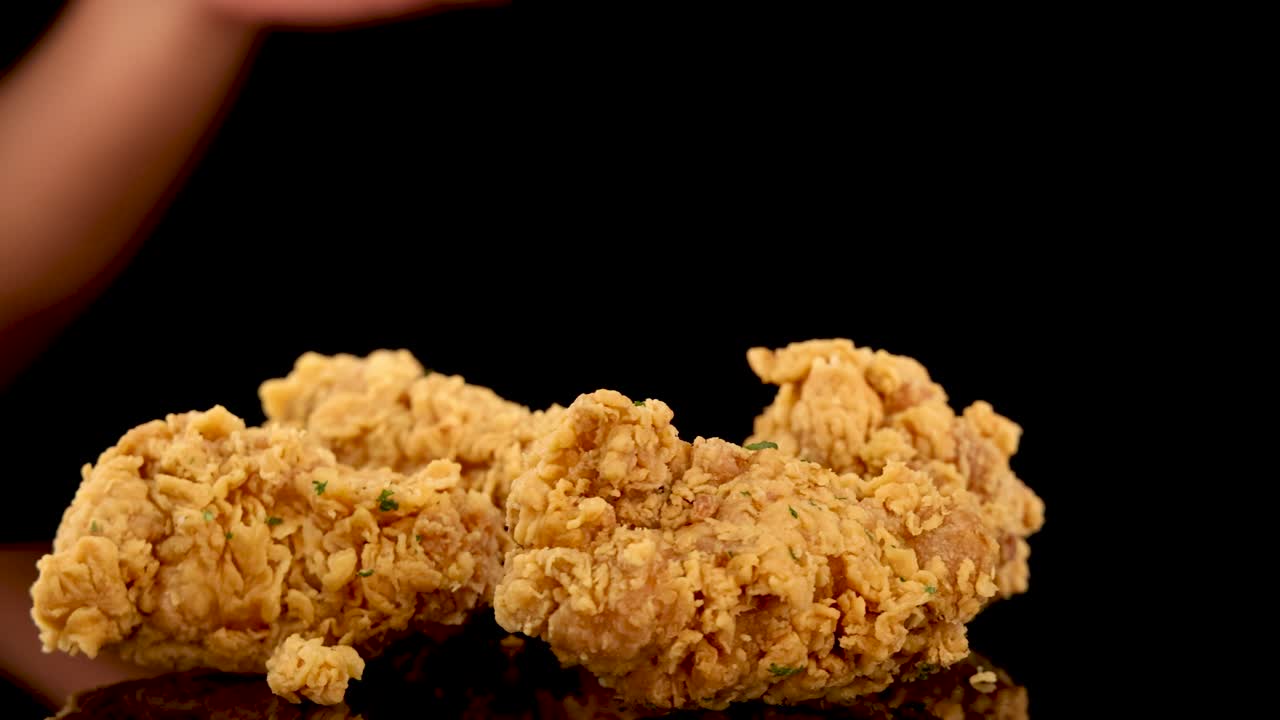A hand reaches in to pick up a piece of golden, crumbed fried chicken from a pile, shot in slow motion against a black background with dramatic lighting