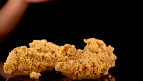 a hand reaches in to pick up a piece of golden, crumbed fried chicken from a pile, shot in slow motion against a black background with dramatic lighting